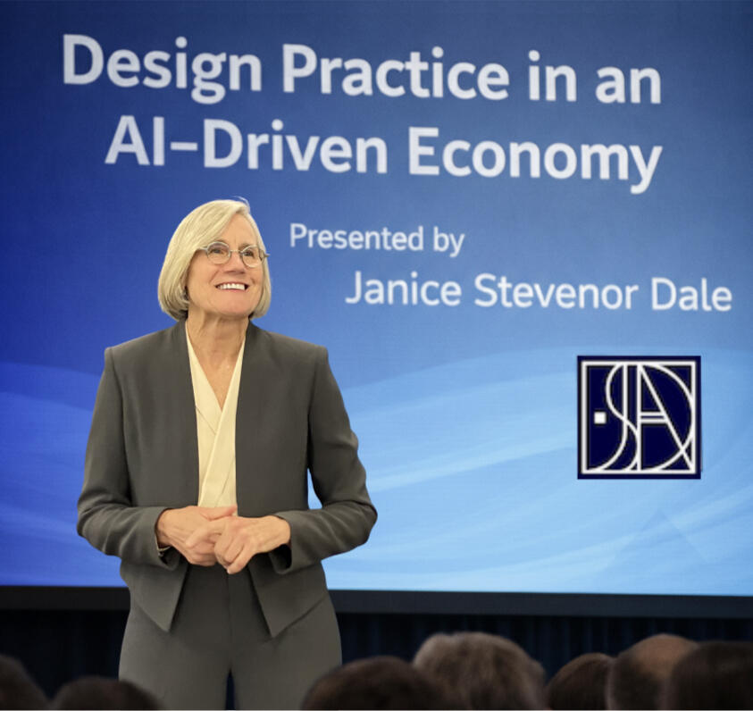 JANICE SPEAKING JANICE SPEAKING IN FRONT OF AN AUDIENCE WITH A VIDEO SCREEN BEHIND HER WITH THE TITLE: "DESIGN PRACTICE IN AN AI-DRIVEN ECONOMY"